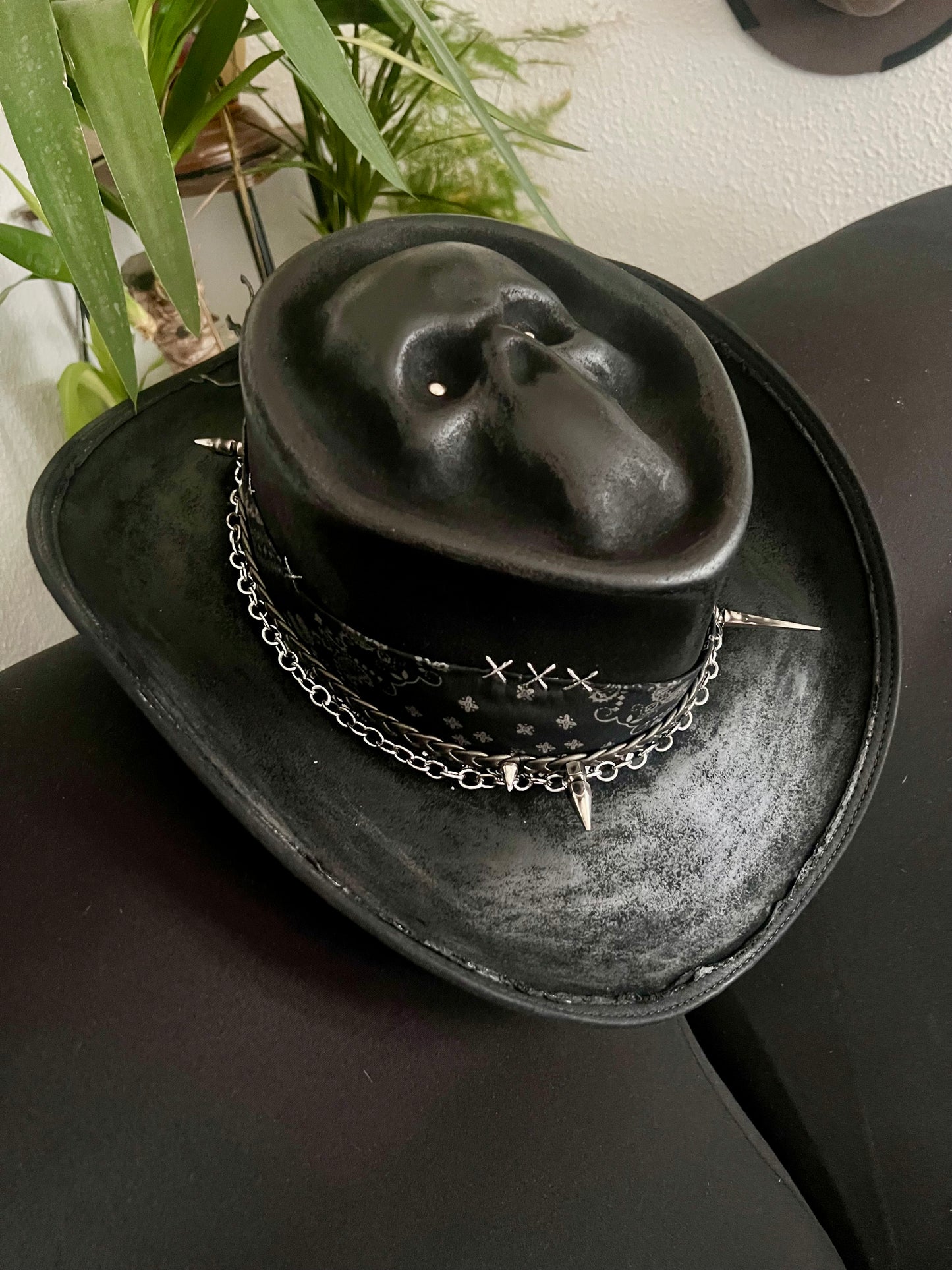 Black skull themed cowboy hat with chains and spikes by JonHats on a black surface