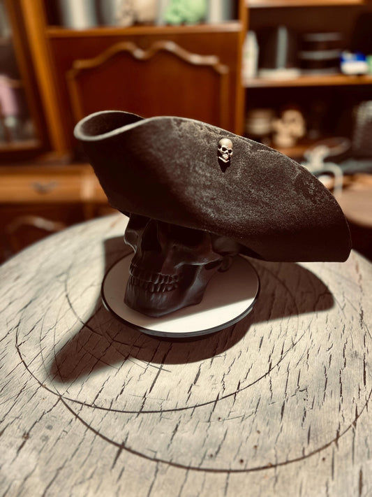 Artisan pirate hat for adults in black with skull pin displayed on skull stand on wooden surface
