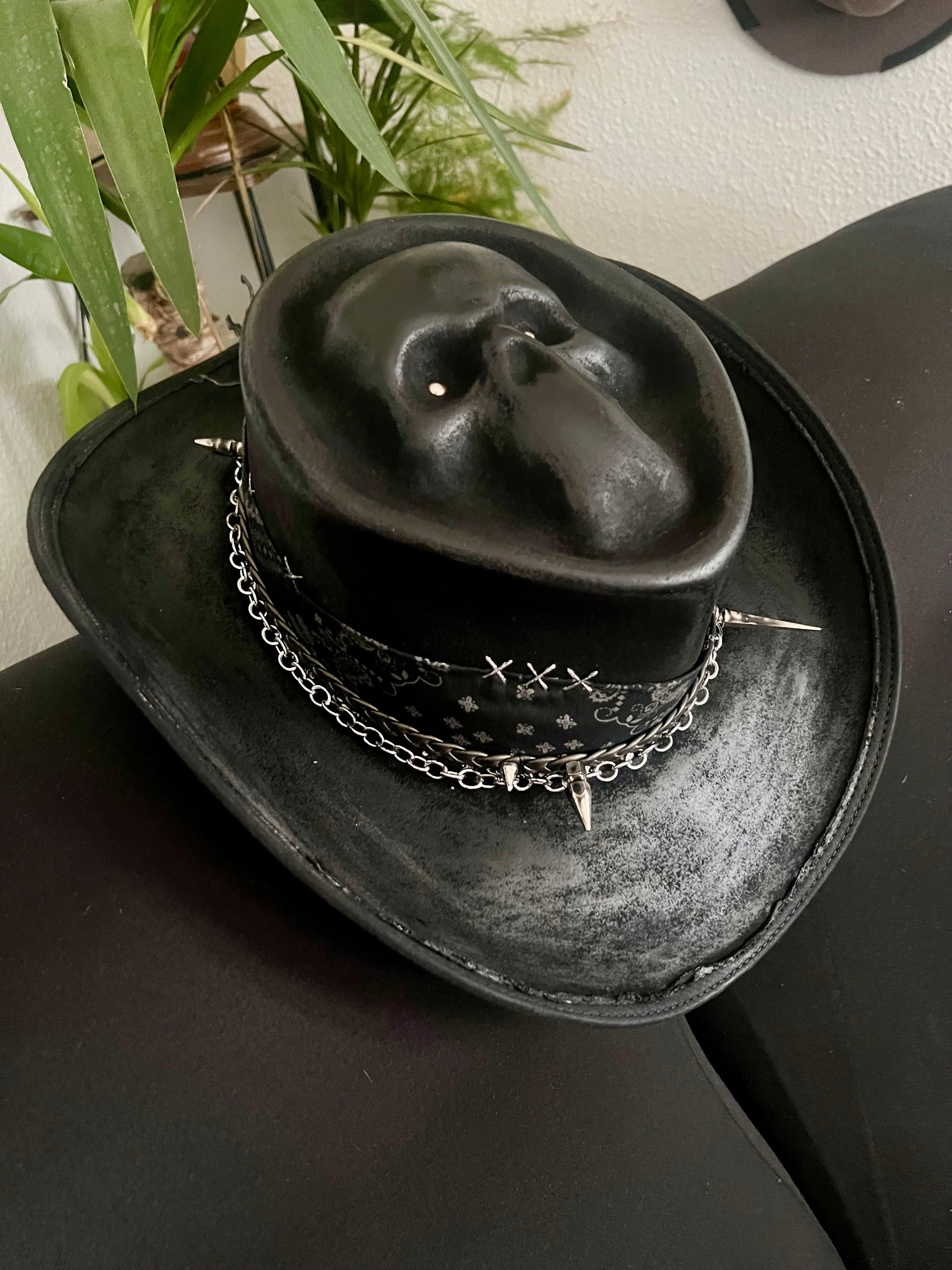 Black skull themed cowboy hat with chains and spikes by JonHats on a black surface